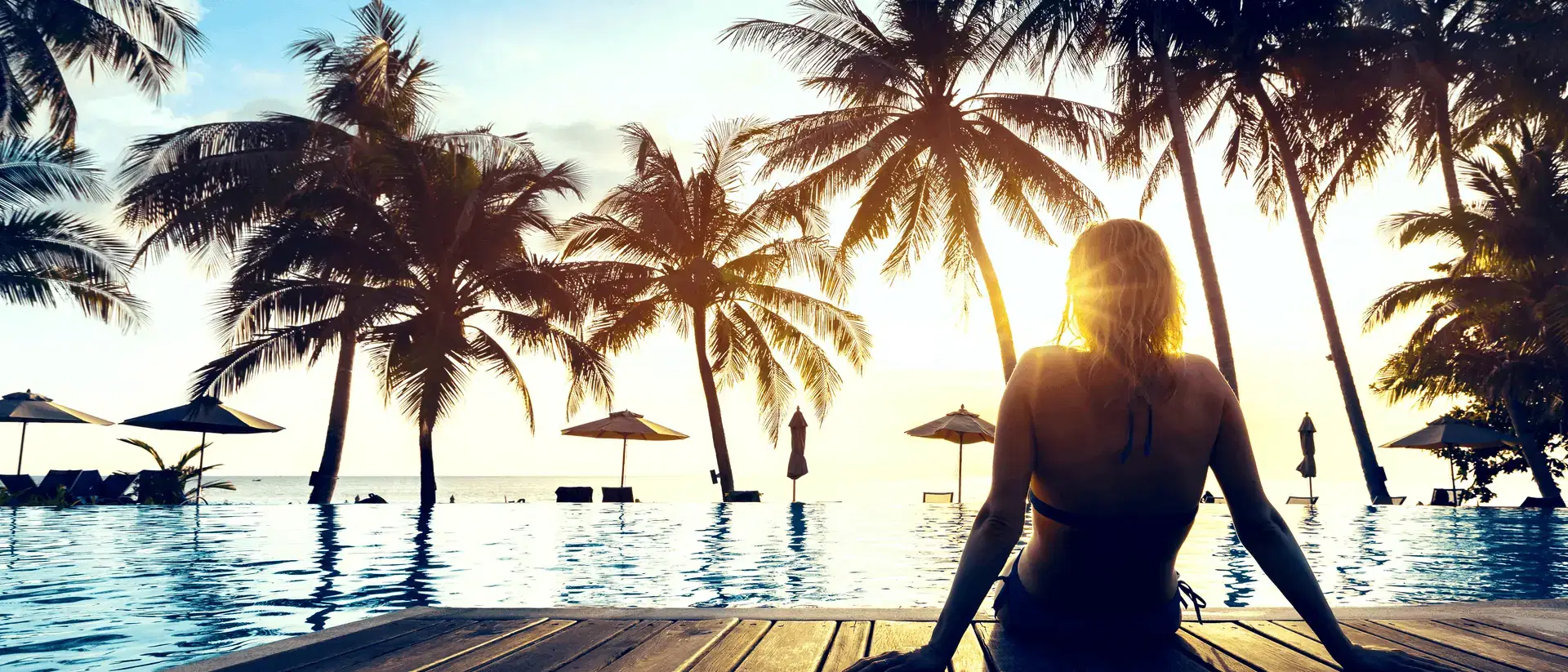 Woman sitting at a tropical infinity pool at sunset, with palm trees and umbrellas over the water