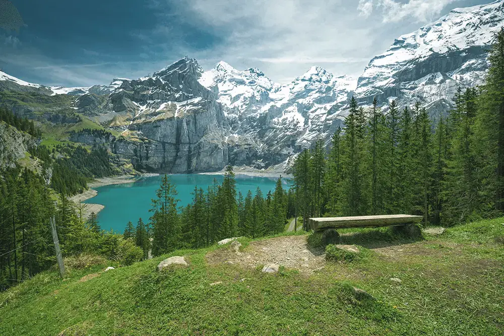 Turquoise alpine lake surrounded by pine forest and snow-capped peaks, with a wooden bench in the foreground